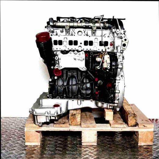 Side view of a diesel engine OM 651.950 on a wooden pallet against a white background. The engine block is black, the cylinder head and oil pan are silver, with yellow markings.